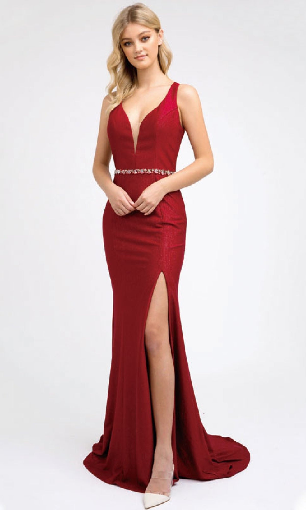 A woman models the Juliet 237 long prom dress, a deep red, form-fitting gown with a plunging neckline, beaded waistband, high slit, strappy back, and floor-length hem as she poses before a plain white background.